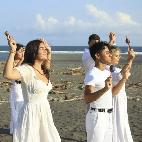 Yoga teacher training students in white holding instruments during a beach ceremony.